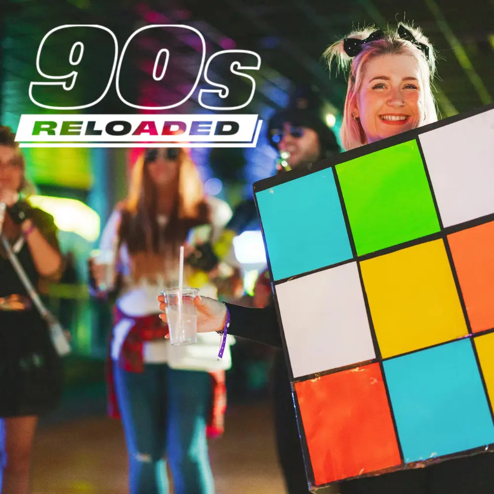 An adult female smiling wearing a rubiks cube costume during a butlins adult weekender with "90's reloaded" displayed in white text