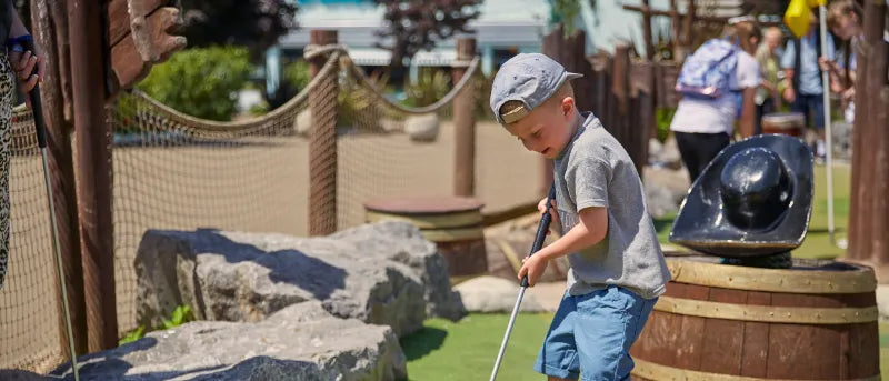 Child playing mini golf at a themed park with props and other visitors in the background.
