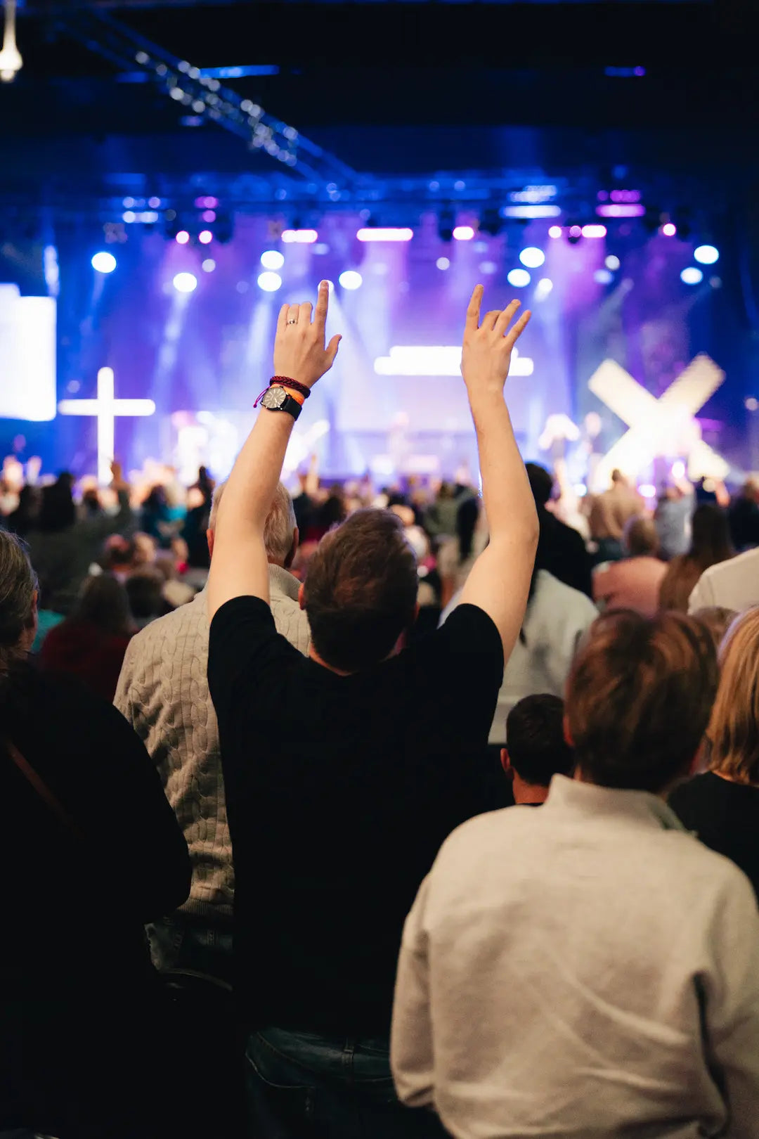 People giving praise with raised hands in a darkly lit auditorium with stage lights at Butlins Minehead Spring Harvest