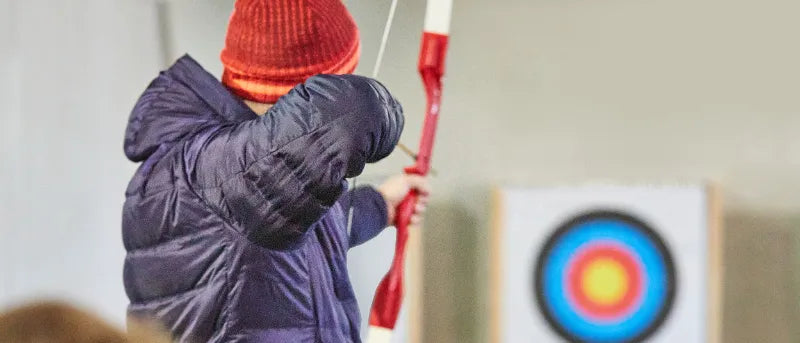 Person in a purple jacket and red beanie holding a bow and arrow with a target in the background